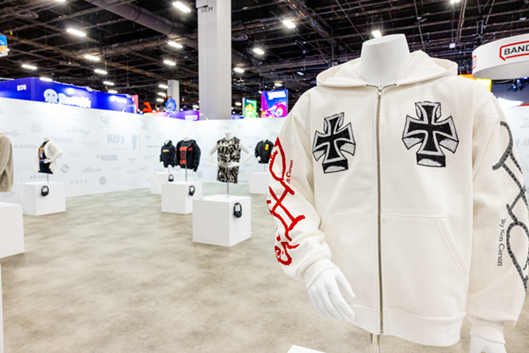 A branded apparel exhibition booth at a licensing trade show, featuring streetwear on mannequins and a white zip-up hoodie with graphic prints in the foreground.