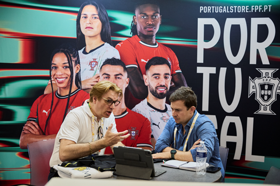 Two business professionals in a meeting at a trade show, seated in front of a large Portugal National Football Team promotional backdrop.